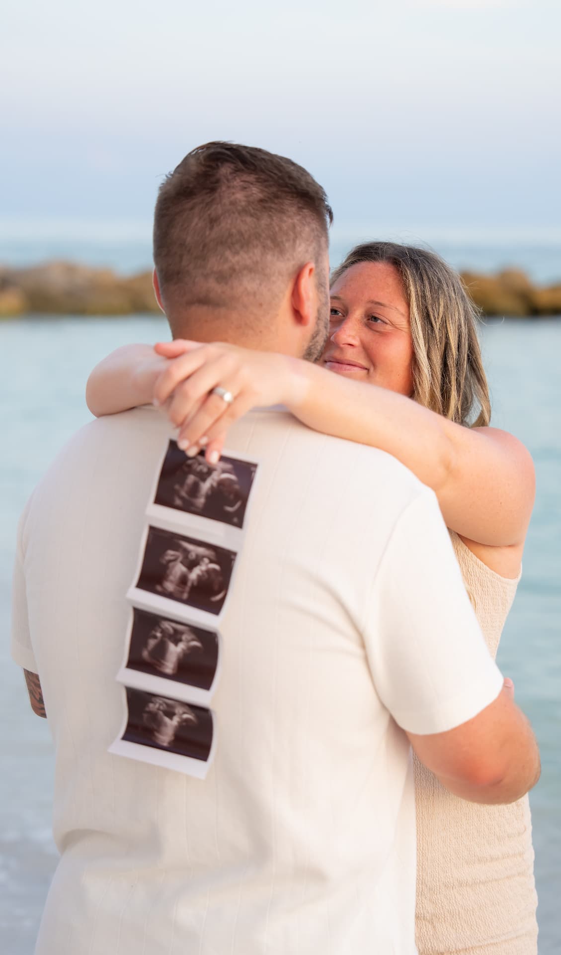Expecting couple embracing on the beach with ultrasound photos held against his back