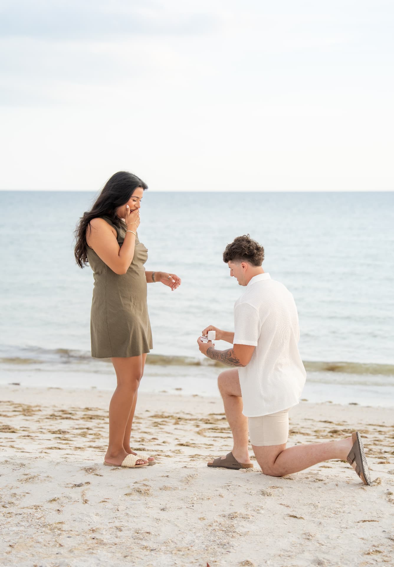 A man on one knee proposing with a ring box on a Marco Island beach as she reacts in surprise