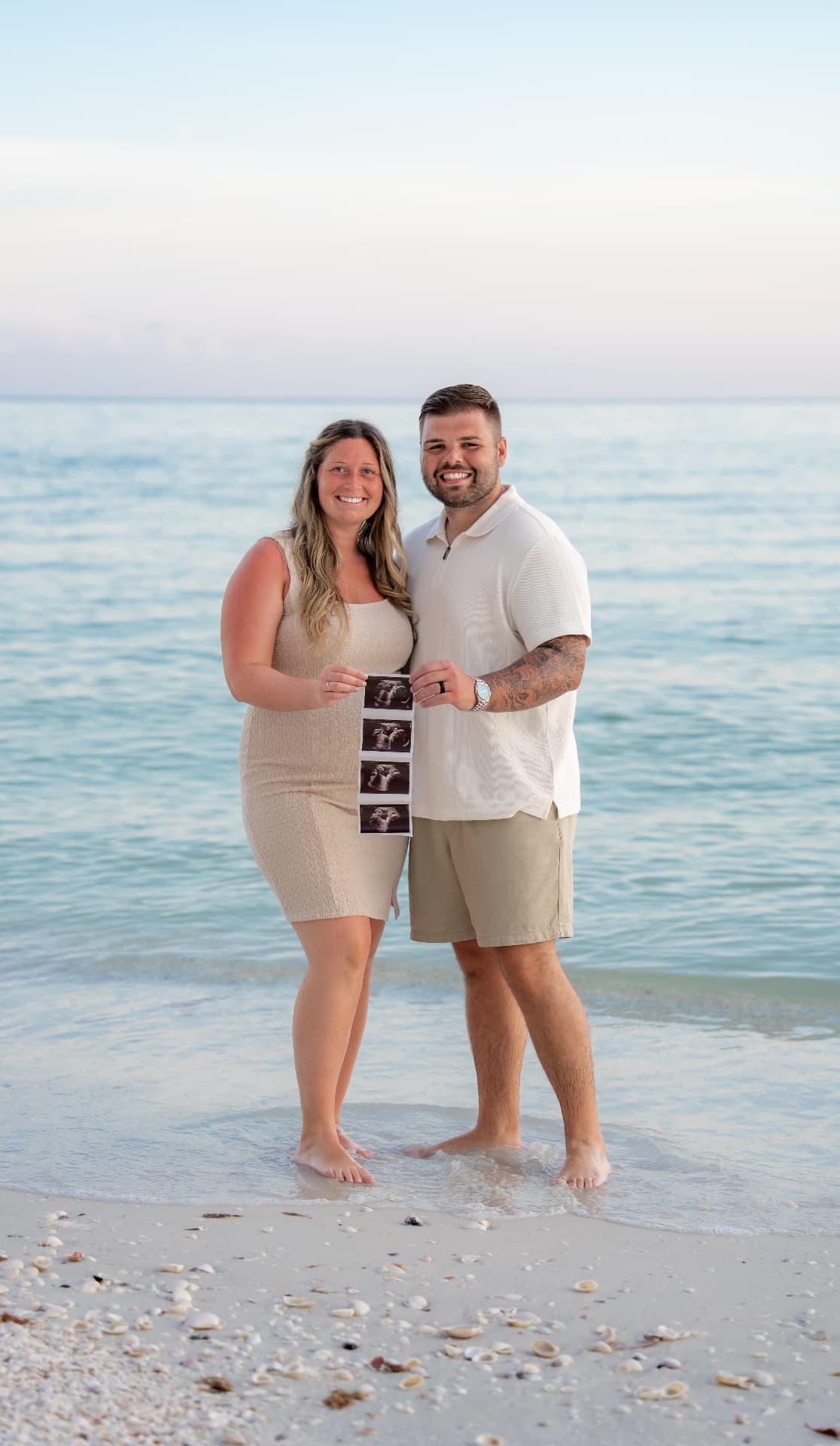 Expecting parents holding up ultrasound photos while standing in the shallow gulf water at sunset