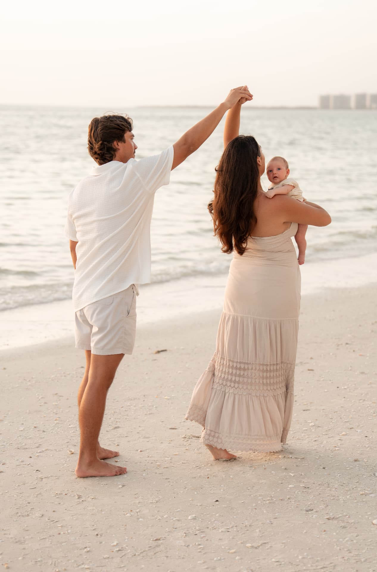 Parents dancing on the sand at sunset while mom holds their baby